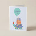 A folded greeting card standing upright on a light beige surface. The card features a whimsical illustration of a turtle wearing a party hat and carrying stacked presents under a mint green balloon that reads 'HAPPY BIRTHDAY PHOEBE'.