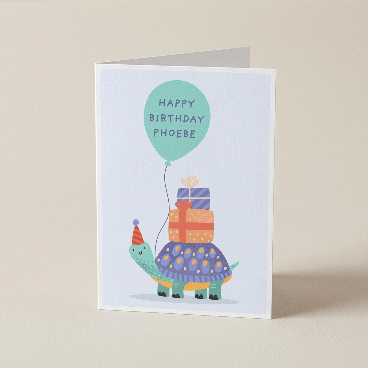 A folded greeting card standing upright on a light beige surface. The card features a whimsical illustration of a turtle wearing a party hat and carrying stacked presents under a mint green balloon that reads 'HAPPY BIRTHDAY PHOEBE'.
