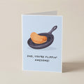 A folded greeting card standing upright on a light beige surface. The card has a pale blue background and features an illustration of a golden-brown pancake in a black skillet, with the text 'DAD, YOU'RE FLIPPIN' AWESOME!' printed below.