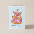 A folded greeting card standing upright on a light beige surface. The card features an illustration of a pink ghost-themed cake with orange frosting and small white ghosts, with the text 'happy boo-thday!' printed below.