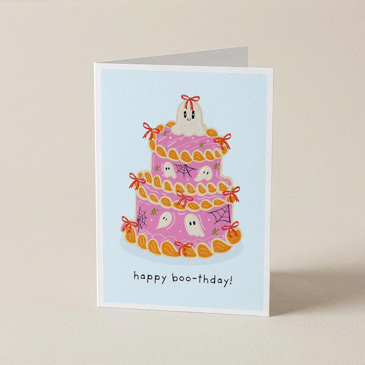 A folded greeting card standing upright on a light beige surface. The card features an illustration of a pink ghost-themed cake with orange frosting and small white ghosts, with the text 'happy boo-thday!' printed below.