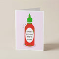 A folded greeting card standing upright on a light beige surface. The card features a stylized illustration of a red hot sauce bottle with a green cap and the text 'HAPPY BIRTHDAY HOT STUFF' on the front.