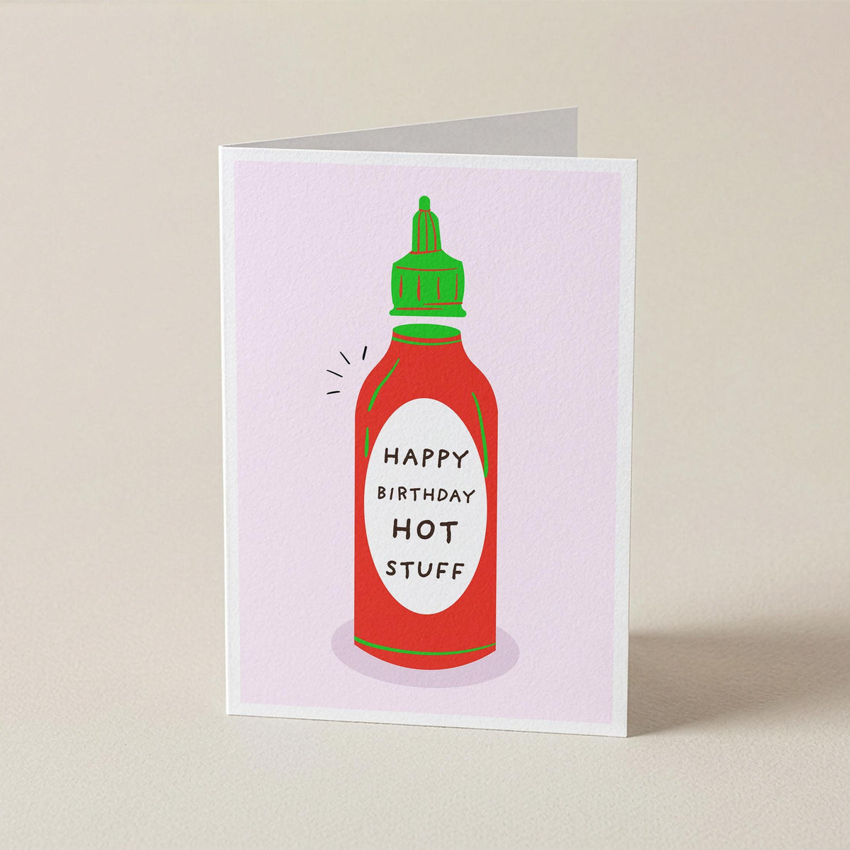 A folded greeting card standing upright on a light beige surface. The card features a stylized illustration of a red hot sauce bottle with a green cap and the text 'HAPPY BIRTHDAY HOT STUFF' on the front.