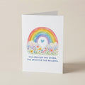 A white greeting card standing upright on a light beige background, featuring a watercolor rainbow, flowers, a small blue heart, and the printed phrase, 'THE GREATER THE STORM, THE BRIGHTER THE RAINBOW.'.