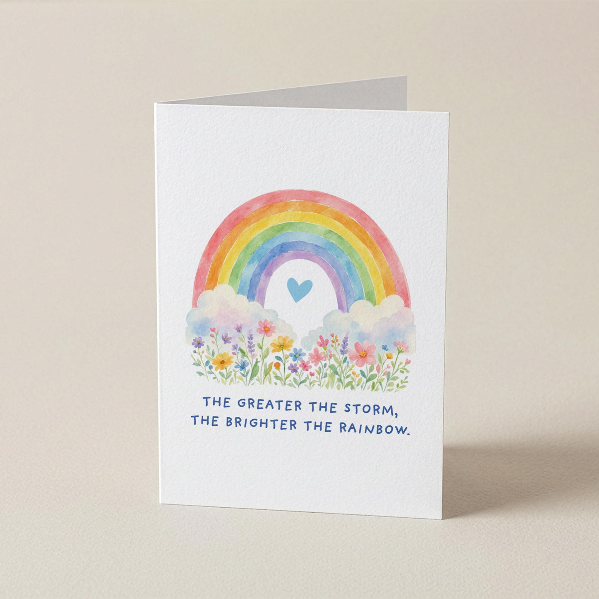 A white greeting card standing upright on a light beige background, featuring a watercolor rainbow, flowers, a small blue heart, and the printed phrase, 'THE GREATER THE STORM, THE BRIGHTER THE RAINBOW.'.