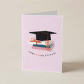 A folded light pink graduation card standing upright on a warm beige background. The front features an illustration of a black graduation cap resting on stacked books with a peach ribbon, and the word 'CONGRADULATIONS!' printed below in black with peach lettering.