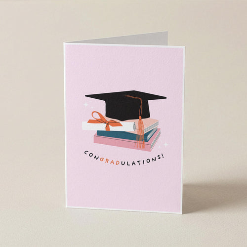 A folded light pink graduation card standing upright on a warm beige background. The front features an illustration of a black graduation cap resting on stacked books with a peach ribbon, and the word 'CONGRADULATIONS!' printed below in black with peach lettering.