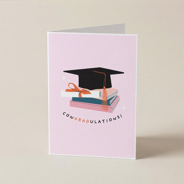 A folded light pink graduation card standing upright on a warm beige background. The front features an illustration of a black graduation cap resting on stacked books with a peach ribbon, and the word 'CONGRADULATIONS!' printed below in black with peach lettering.