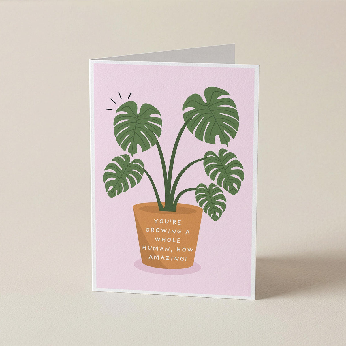 A folded greeting card standing upright on a light beige surface. The card has a pale pink background with an illustration of a potted monstera plant and the text, 'YOU'RE GROWING A WHOLE HUMAN, NOW AMAZING!' in a terracotta pot.