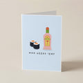 A folded greeting card standing upright on a beige surface. The card has a pale blue front with an illustration of two sushi rolls and a pink bottle labeled 'MANGAJOY', above the text 'WHO NEEDS "EM?'.