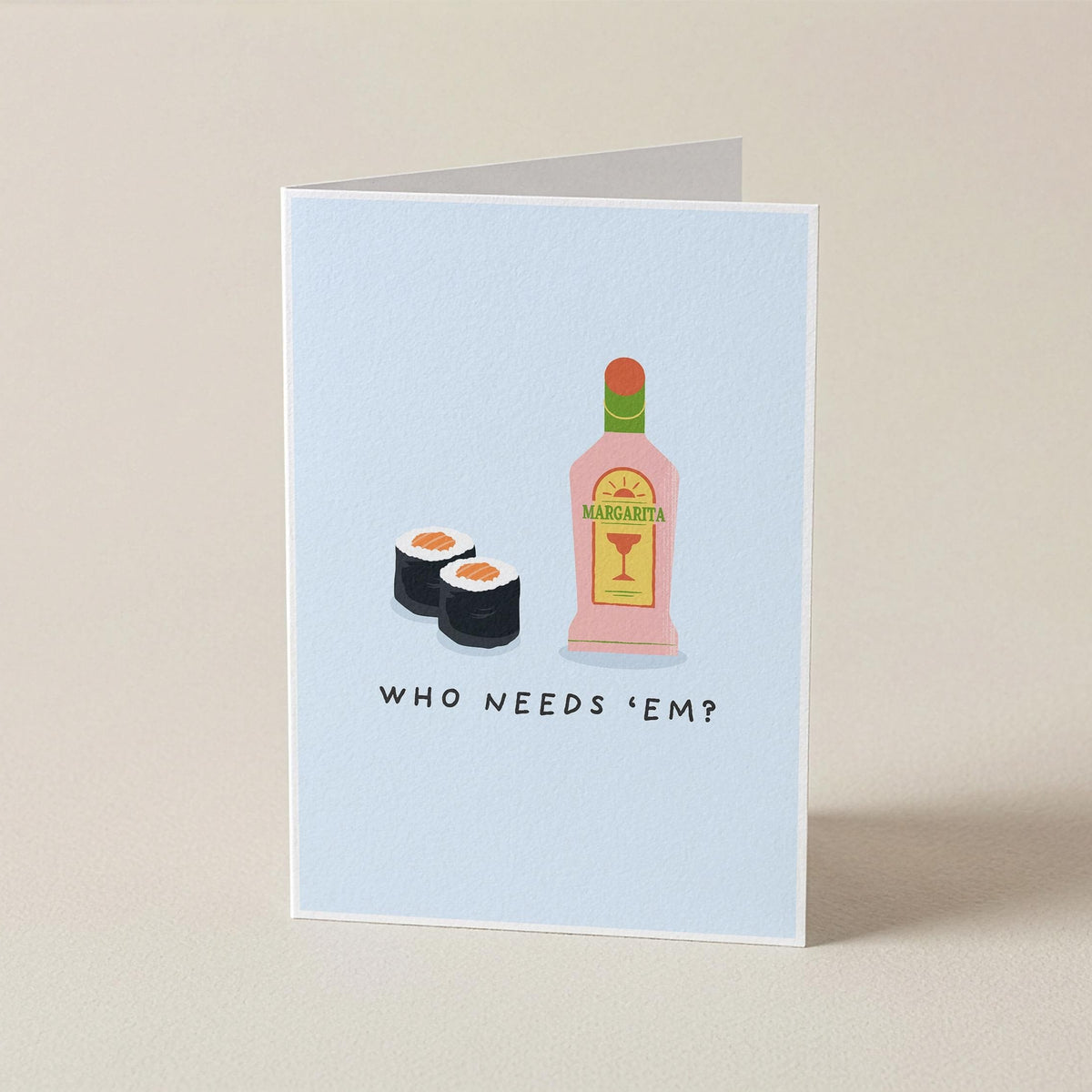A folded greeting card standing upright on a beige surface. The card has a pale blue front with an illustration of two sushi rolls and a pink bottle labeled 'MANGAJOY', above the text 'WHO NEEDS "EM?'.