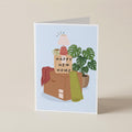 A folded greeting card standing upright on a light beige surface. The card features an illustration of a cardboard box with a sign reading 'HAPPY NEW HOME,' a pink lamp, a green plant, and folded blankets in a soft pastel palette.
