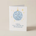 A white greeting card standing upright on a beige surface, featuring a blue mirrored disco ball, small gold stars, and the text 'SARAH, WILL YOU BE MY BRIDESMAID?'.
