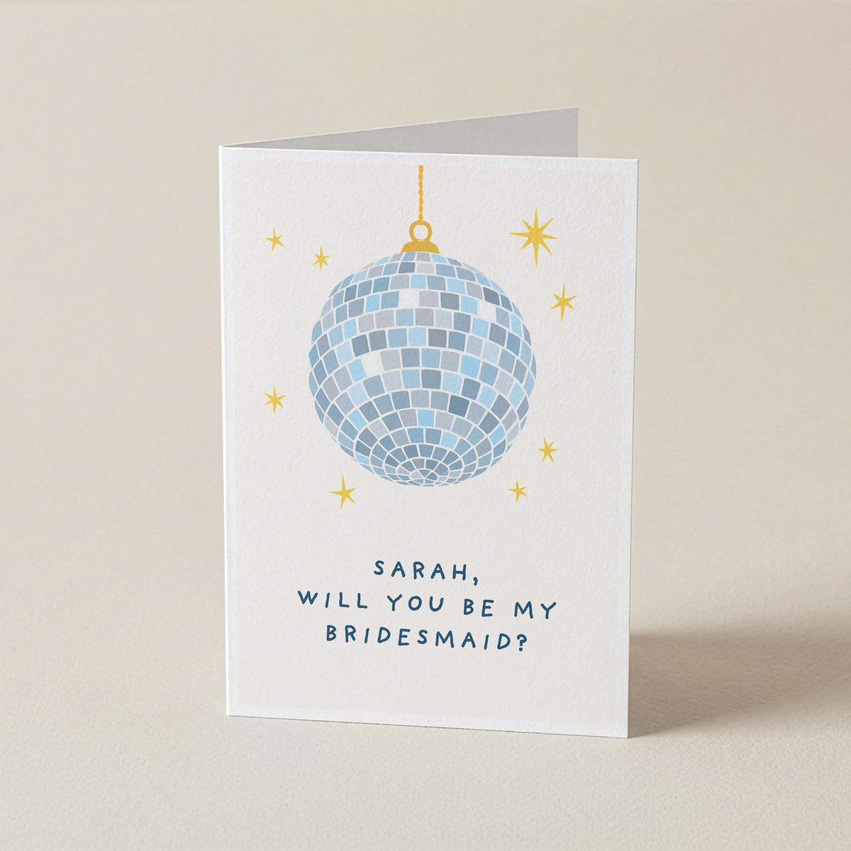 A white greeting card standing upright on a beige surface, featuring a blue mirrored disco ball, small gold stars, and the text 'SARAH, WILL YOU BE MY BRIDESMAID?'.