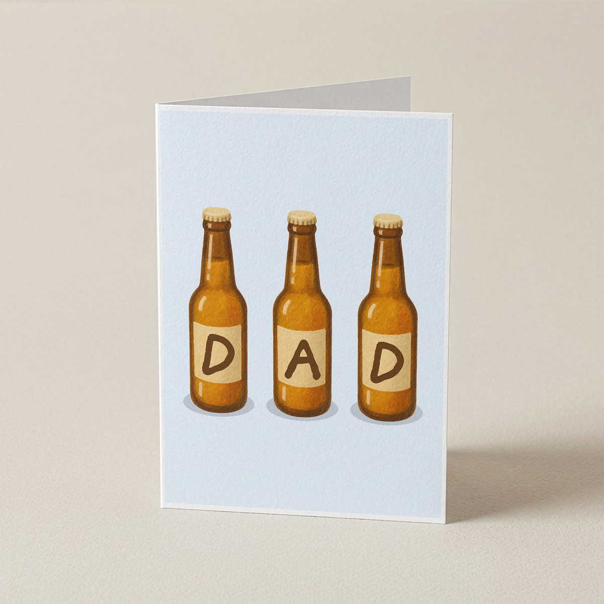 A folded greeting card standing upright on a light beige surface. The card has a pale blue front featuring an illustration of three brown beer bottles with cream labels reading 'D', 'A', and 'D' to spell 'DAD'.