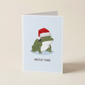 A folded greeting card standing upright on a light beige surface. The card features an illustration of a green frog wearing a red Santa hat on a pale blue background, with the text 'MISTLE-TOAD.