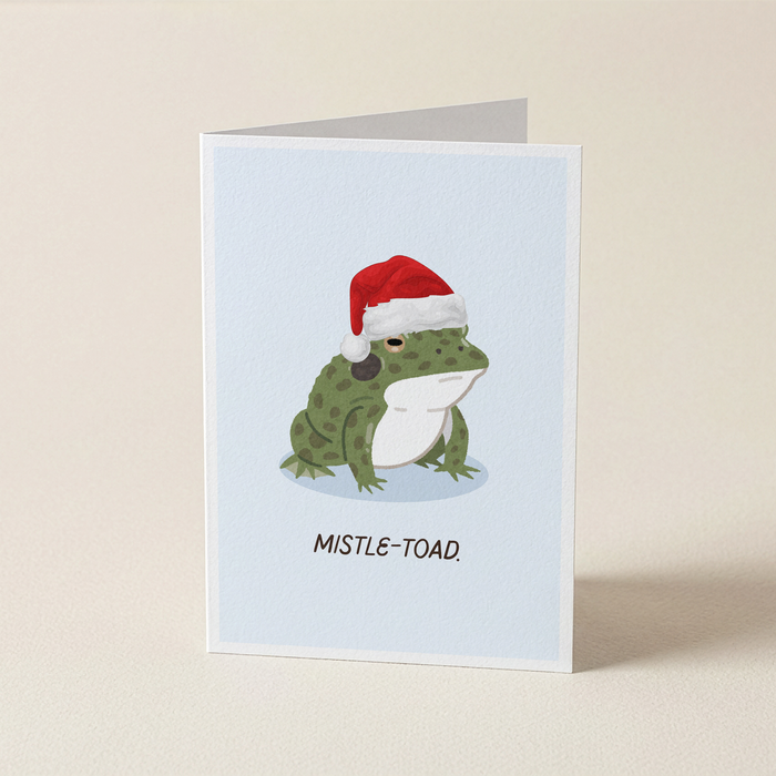 A folded greeting card standing upright on a light beige surface. The card features an illustration of a green frog wearing a red Santa hat on a pale blue background, with the text 'MISTLE-TOAD.