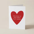 A white greeting card standing upright on a beige surface, featuring a large red heart with the text 'WELCOME TO THE WORLD TINY HUMAN' in white lettering.