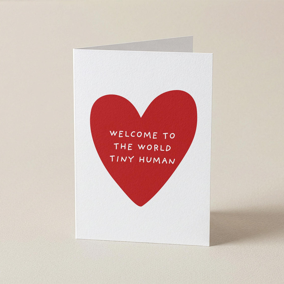 A white greeting card standing upright on a beige surface, featuring a large red heart with the text 'WELCOME TO THE WORLD TINY HUMAN' in white lettering.