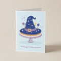 A folded greeting card standing upright on a beige surface. The card features an illustration of a blue witch hat with stars and wand toppers on a cake stand, with the text 'sending birthday witches!' printed below.
