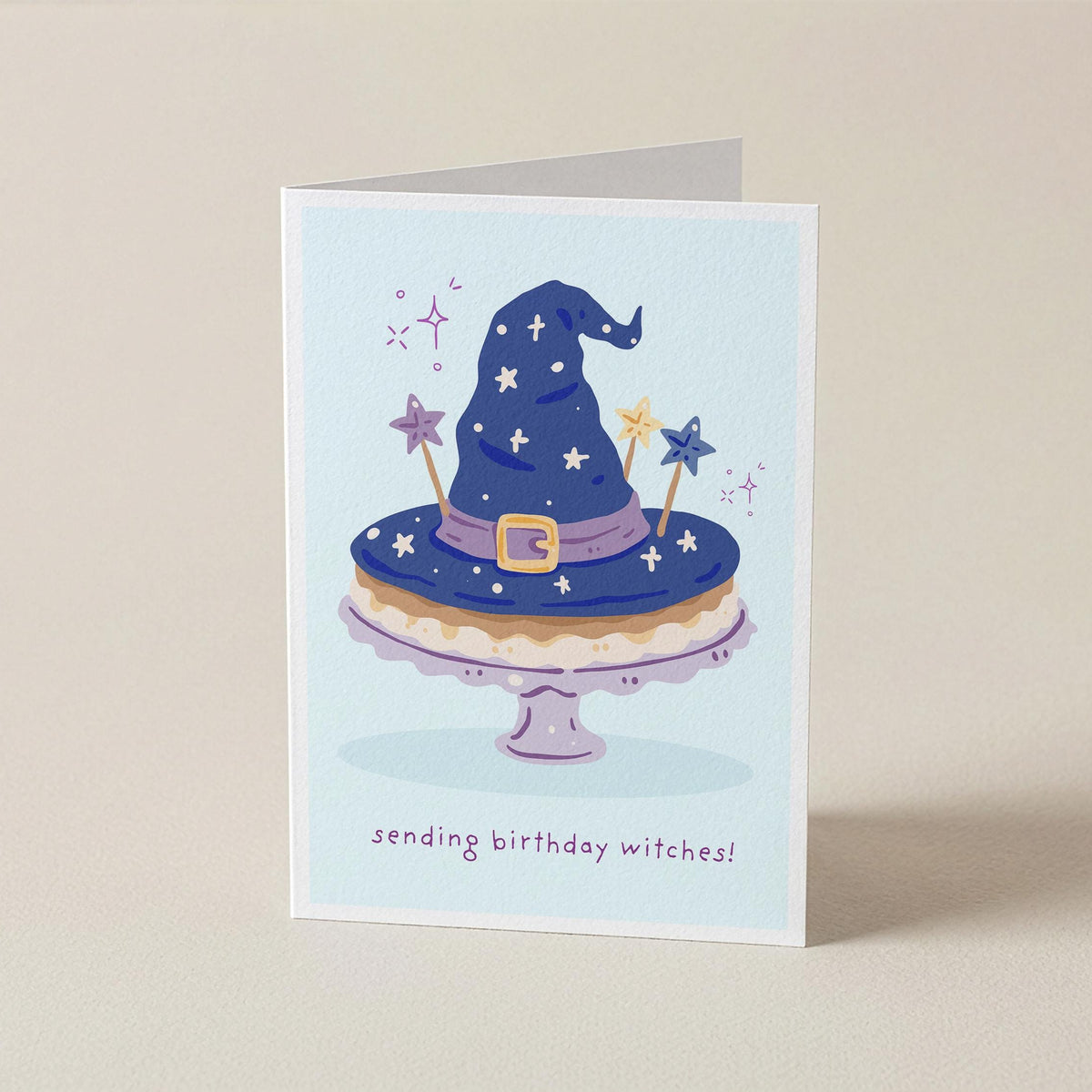 A folded greeting card standing upright on a beige surface. The card features an illustration of a blue witch hat with stars and wand toppers on a cake stand, with the text 'sending birthday witches!' printed below.