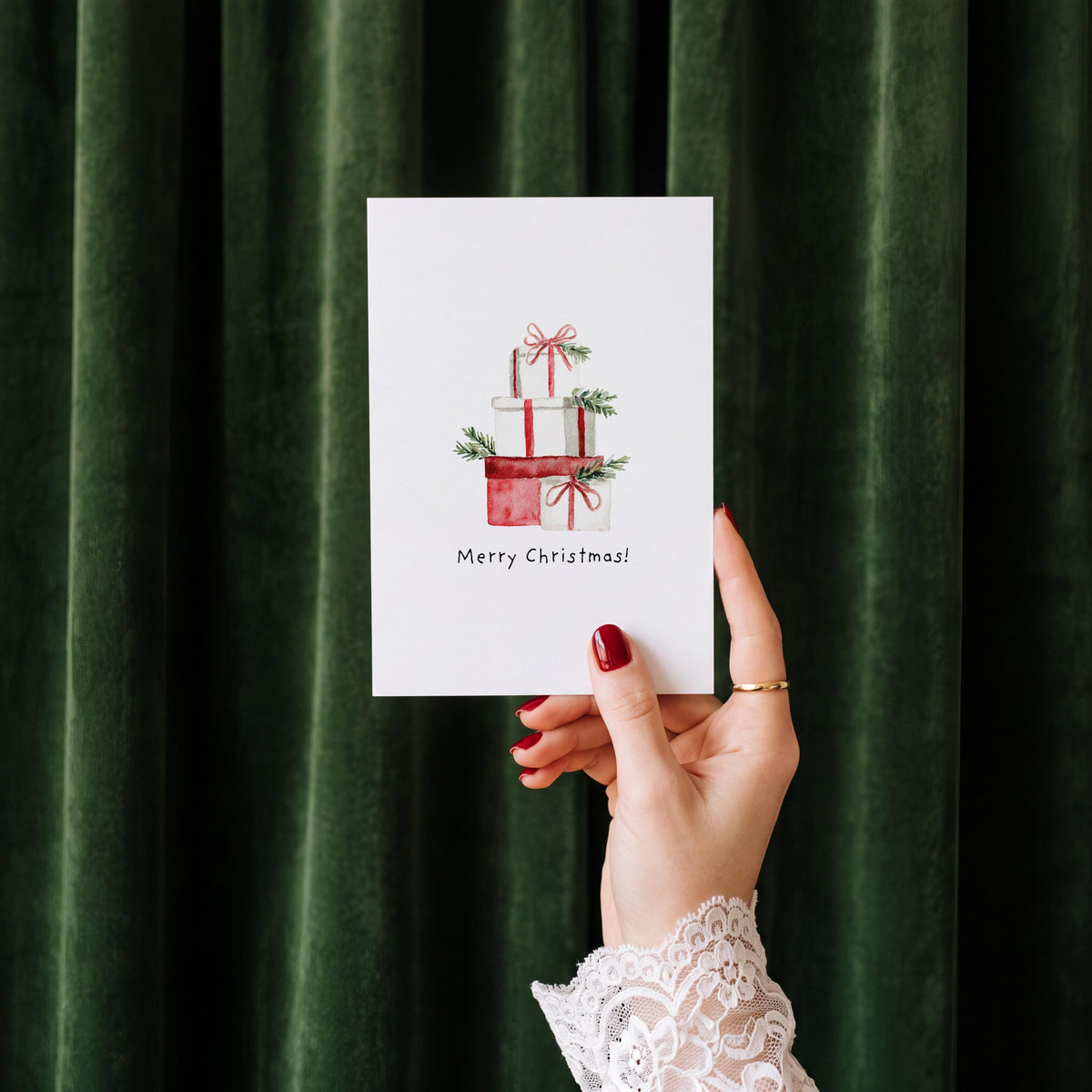 A hand with red nail polish and a white lace cuff holds a white Christmas greeting card featuring a watercolor illustration of stacked red gift boxes with greenery and the text 'Merry Christmas!' against a dark green velvet curtain background.