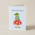 A folded greeting card standing upright on a light beige surface. The card has a white border and features a cute illustration of a green caterpillar wearing a red-and-white polka dot mushroom cap and green leafy hat, holding a small gift box.