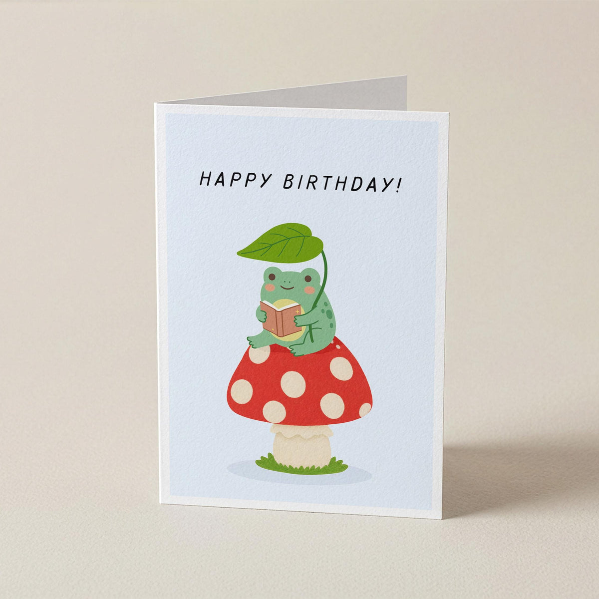 A folded greeting card standing upright on a light beige surface. The card has a white border and features a cute illustration of a green caterpillar wearing a red-and-white polka dot mushroom cap and green leafy hat, holding a small gift box.