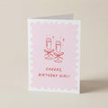 A folded greeting card standing upright on a light beige surface. The card has a white border and a pale pink scalloped center with a red line drawing of two champagne glasses tied with bows and the text 'CHEERS, BIRTHDAY GIRL!'.