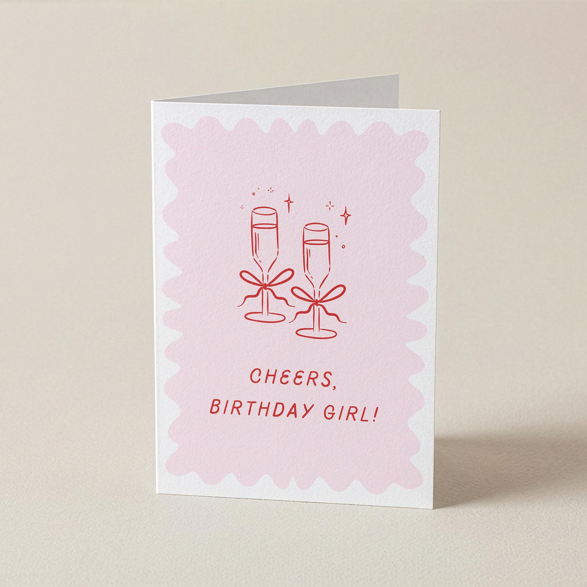 A folded greeting card standing upright on a light beige surface. The card has a white border and a pale pink scalloped center with a red line drawing of two champagne glasses tied with bows and the text 'CHEERS, BIRTHDAY GIRL!'.