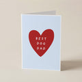 A folded greeting card standing upright on a beige surface. The card has a pale blue background with a large red heart in the center containing the white text 'BEST DOG DAD'.