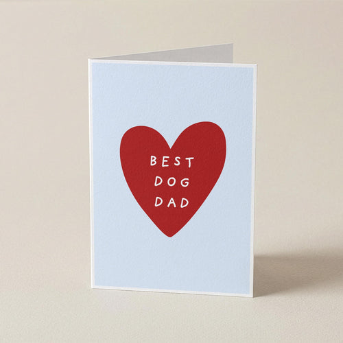 A folded greeting card standing upright on a beige surface. The card has a pale blue background with a large red heart in the center containing the white text 'BEST DOG DAD'.