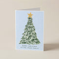 A folded Christmas greeting card standing upright on a beige surface. The card features a watercolor-style green Christmas tree with a gold star on a pale blue background, with the text 'MERRY CHRISTMAS TO THE JONES FAMILY' printed near the bottom.