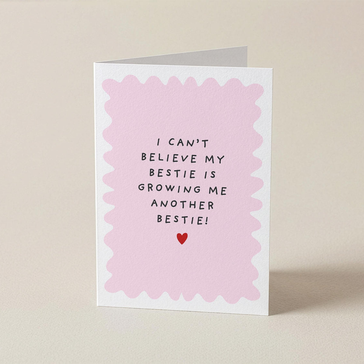 A folded greeting card standing upright on a light beige background. The card has a white border with a scalloped light pink inner frame and black handwritten-style text that reads, 'I CAN'T BELIEVE MY BESTIE IS GROWING ME ANOTHER BESTIE!' with a small red heart below.