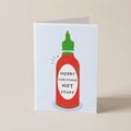 A folded greeting card standing upright on a beige surface, featuring a stylized red hot sauce bottle with a green cap and the text 'MERRY CHRISTMAS HOT STUFF' on the front.