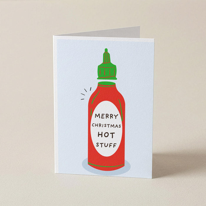 A folded greeting card standing upright on a beige surface, featuring a stylized red hot sauce bottle with a green cap and the text 'MERRY CHRISTMAS HOT STUFF' on the front.