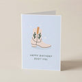 A folded greeting card standing upright on a light beige surface. The card has a pale blue front with an illustration of a cowboy boot in beige and green, above the text 'HAPPY BIRTHDAY BOOT-IFUL'.