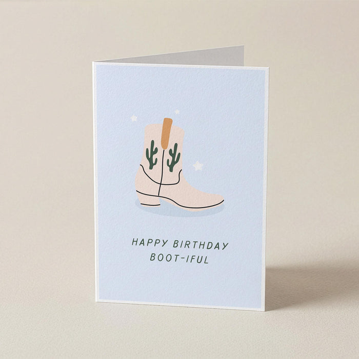 A folded greeting card standing upright on a light beige surface. The card has a pale blue front with an illustration of a cowboy boot in beige and green, above the text 'HAPPY BIRTHDAY BOOT-IFUL'.