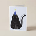 A folded greeting card standing upright on a light beige surface. The card features a minimalist illustration of a black cat wearing a blue party hat with a yellow star, next to the text 'HAPPY BIRTHDAY!'.