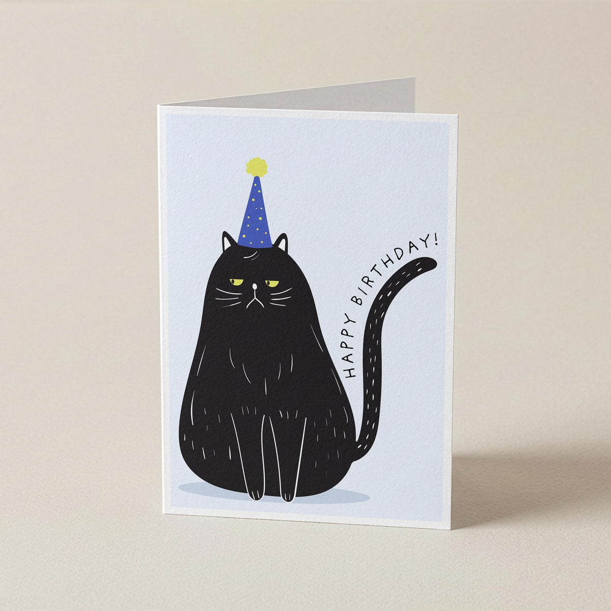 A folded greeting card standing upright on a light beige surface. The card features a minimalist illustration of a black cat wearing a blue party hat with a yellow star, next to the text 'HAPPY BIRTHDAY!'.