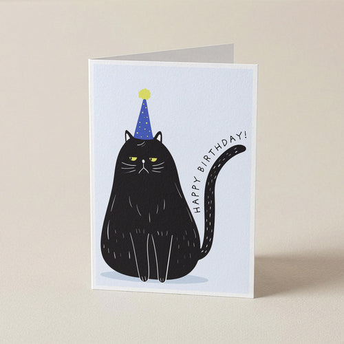 A folded greeting card standing upright on a light beige surface. The card features a minimalist illustration of a black cat wearing a blue party hat with a yellow star, next to the text 'HAPPY BIRTHDAY!'.