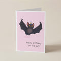 A folded greeting card standing upright on a light beige background. The card has a pink front with an illustration of a dark bat wearing a party hat and the text 'happy birthday you old bat!'.