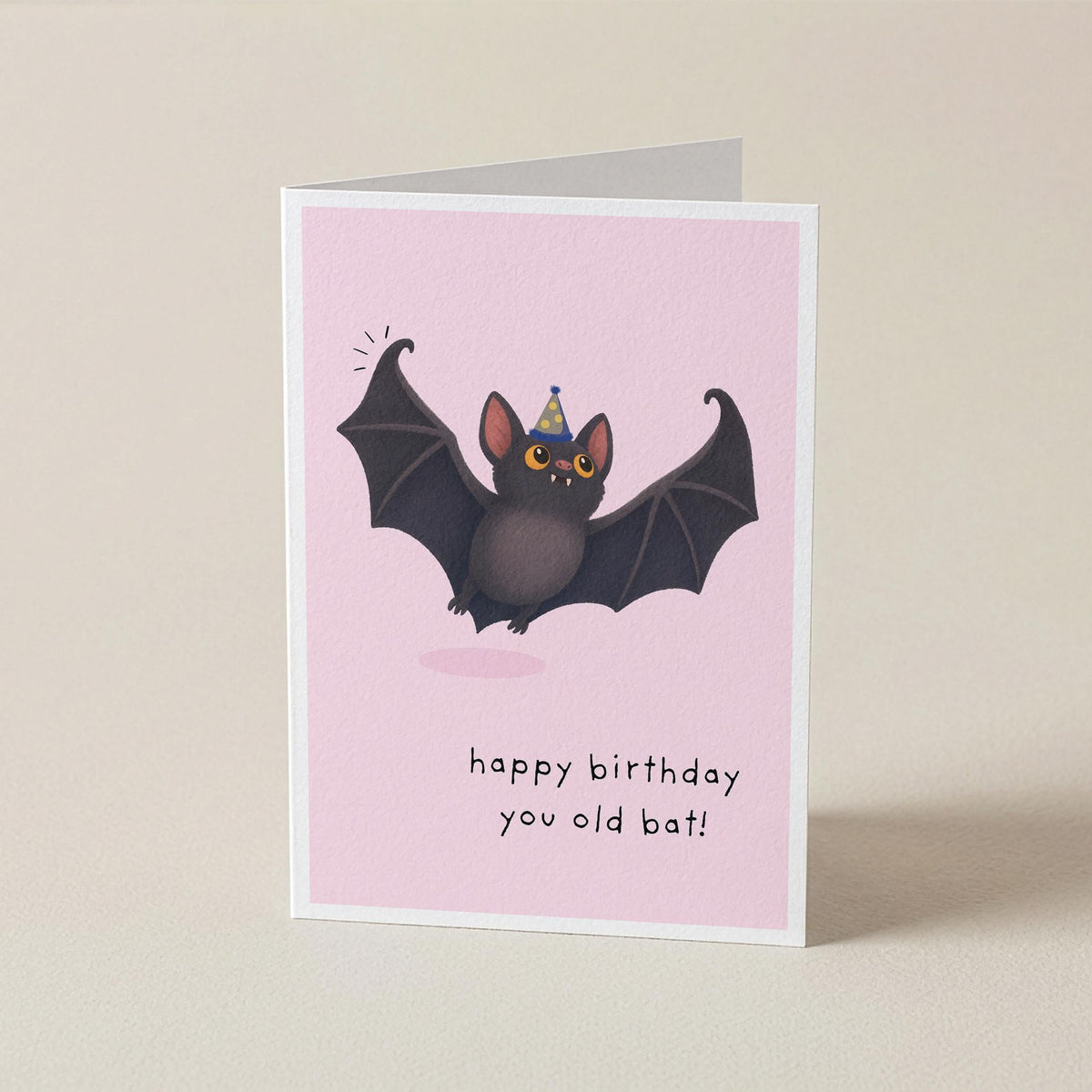 A folded greeting card standing upright on a light beige background. The card has a pink front with an illustration of a dark bat wearing a party hat and the text 'happy birthday you old bat!'.