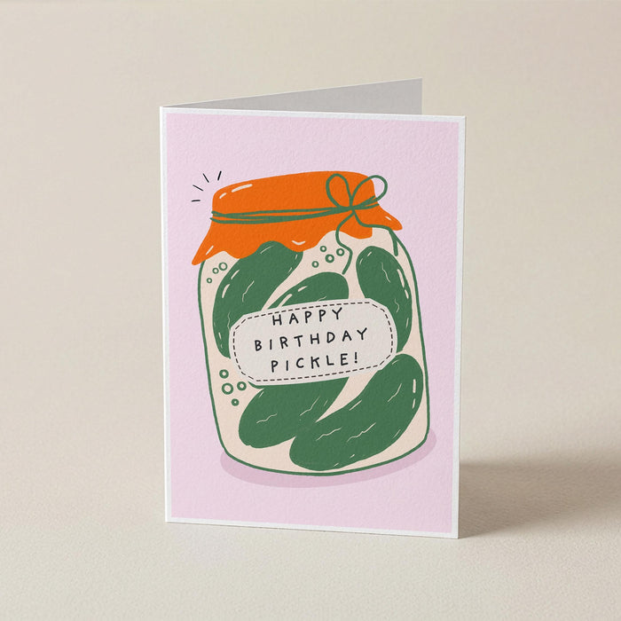 A folded greeting card standing upright on a light beige surface. The card has a pale pink background and features an illustration of a glass jar filled with green pickles, topped with an orange lid or cloth, with a label that reads “HAPPY BIRTHDAY PICKLE!”.