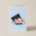 A folded greeting card standing upright on a light beige surface. The card has a pale blue front with an illustration of stacked books, a black graduation cap reading 'CON-GRADS!', and a rolled diploma tied with an orange ribbon.