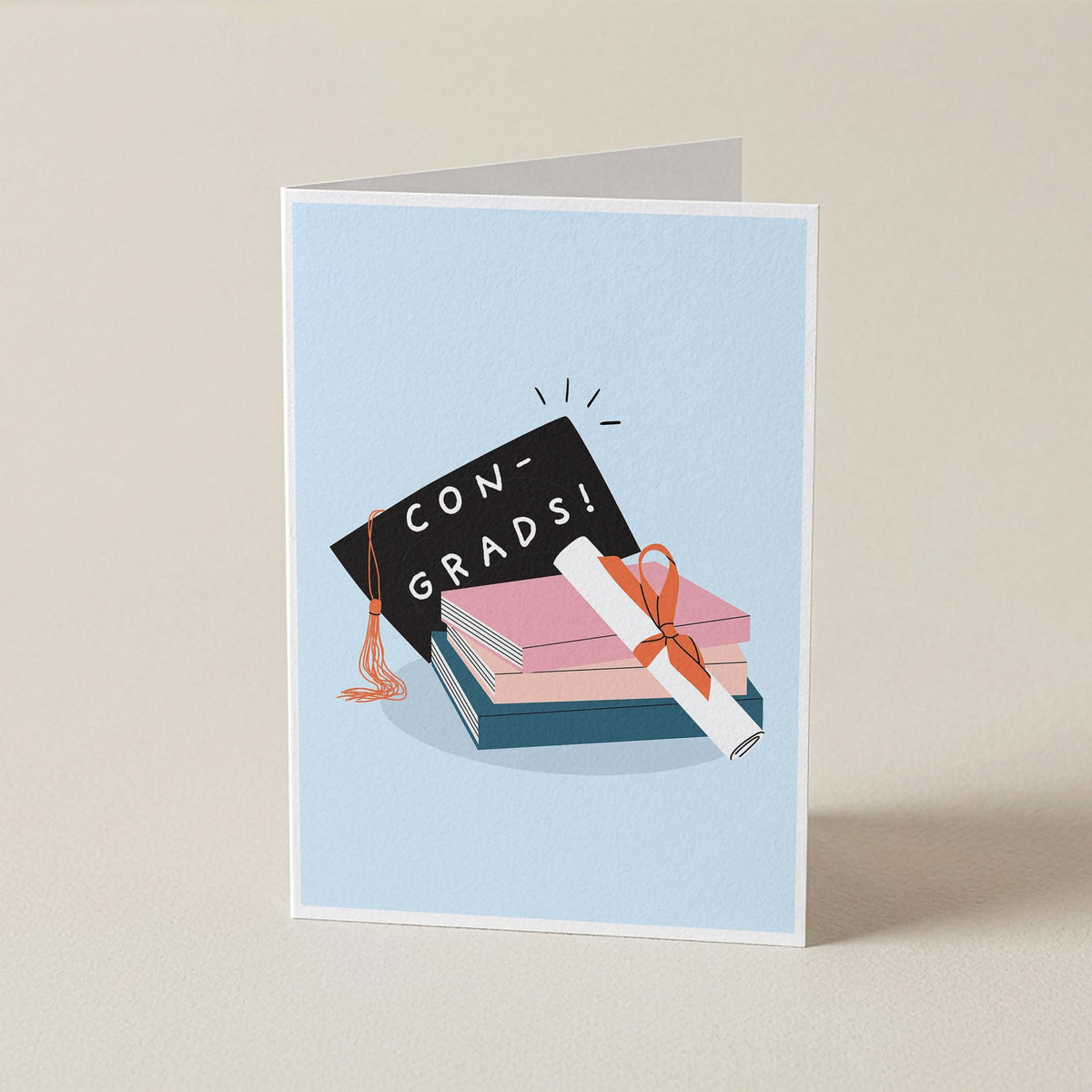 A folded greeting card standing upright on a light beige surface. The card has a pale blue front with an illustration of stacked books, a black graduation cap reading 'CON-GRADS!', and a rolled diploma tied with an orange ribbon.