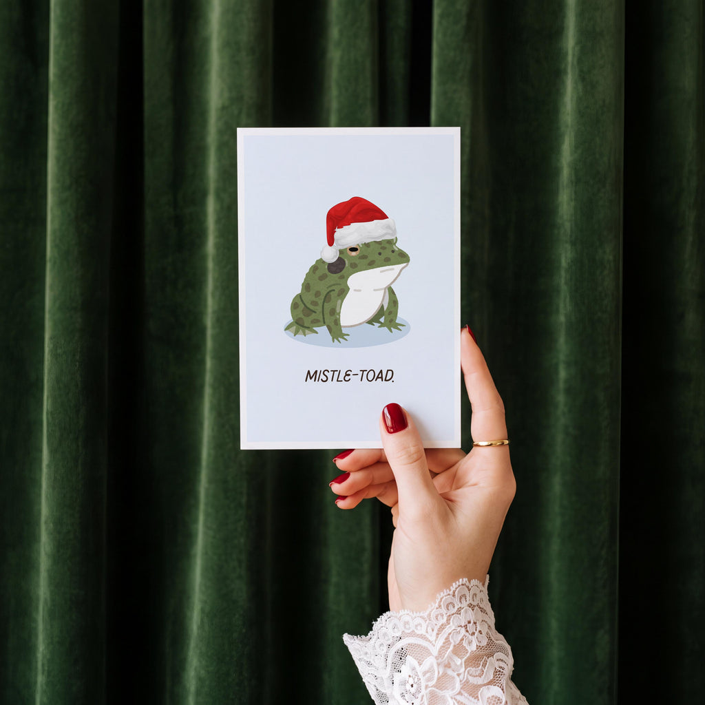 A hand with red nail polish and a white lace sleeve holds up a white greeting card featuring an illustration of a green mistle-toad wearing a red Santa hat, set against a dark green velvet curtain background.