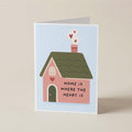 A folded greeting card standing upright on a light beige surface. The card features a cute illustration of a pink house with a green roof, hearts coming from the chimney, and the text 'HOME IS WHERE THE HEART IS' on the front.