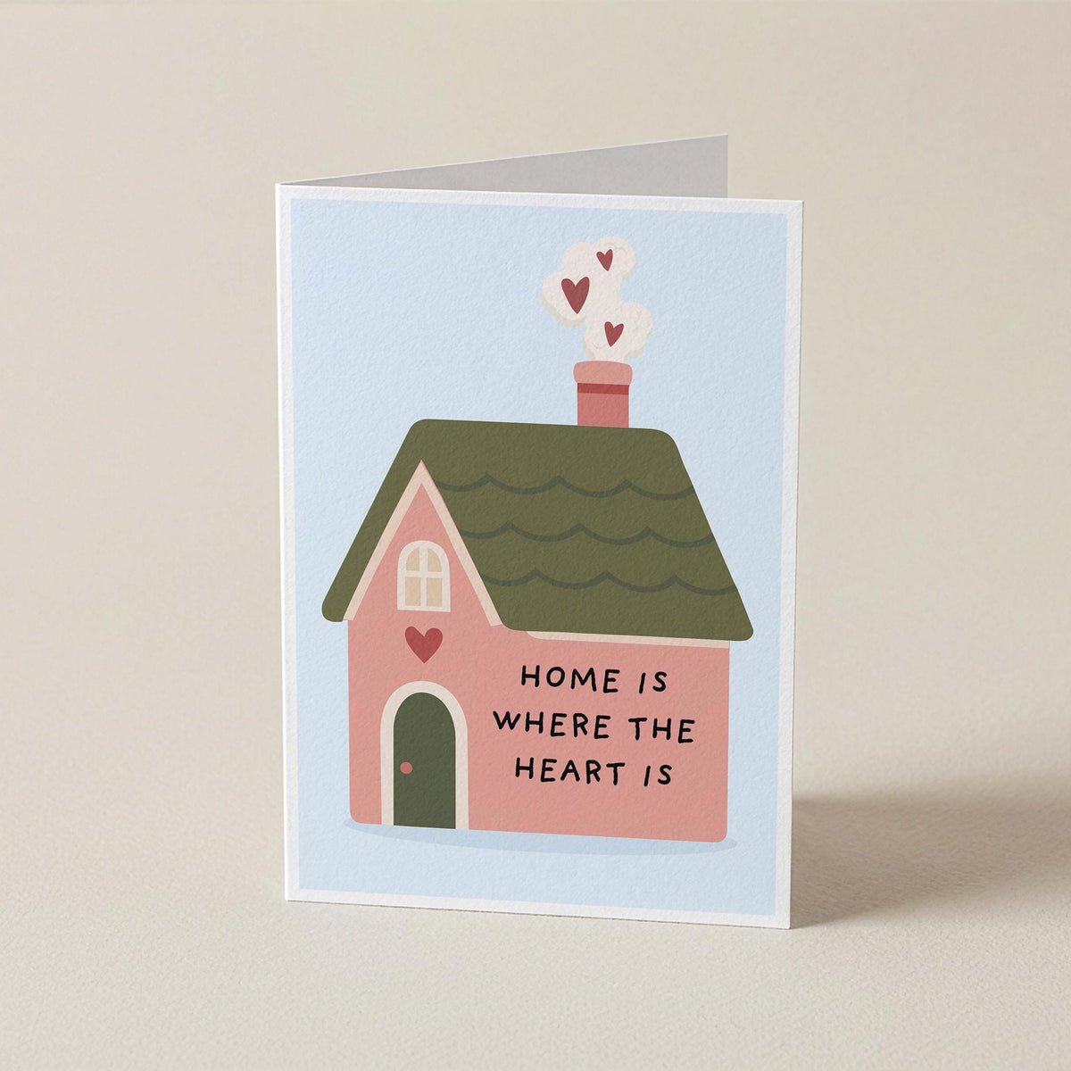 A folded greeting card standing upright on a light beige surface. The card features a cute illustration of a pink house with a green roof, hearts coming from the chimney, and the text 'HOME IS WHERE THE HEART IS' on the front.