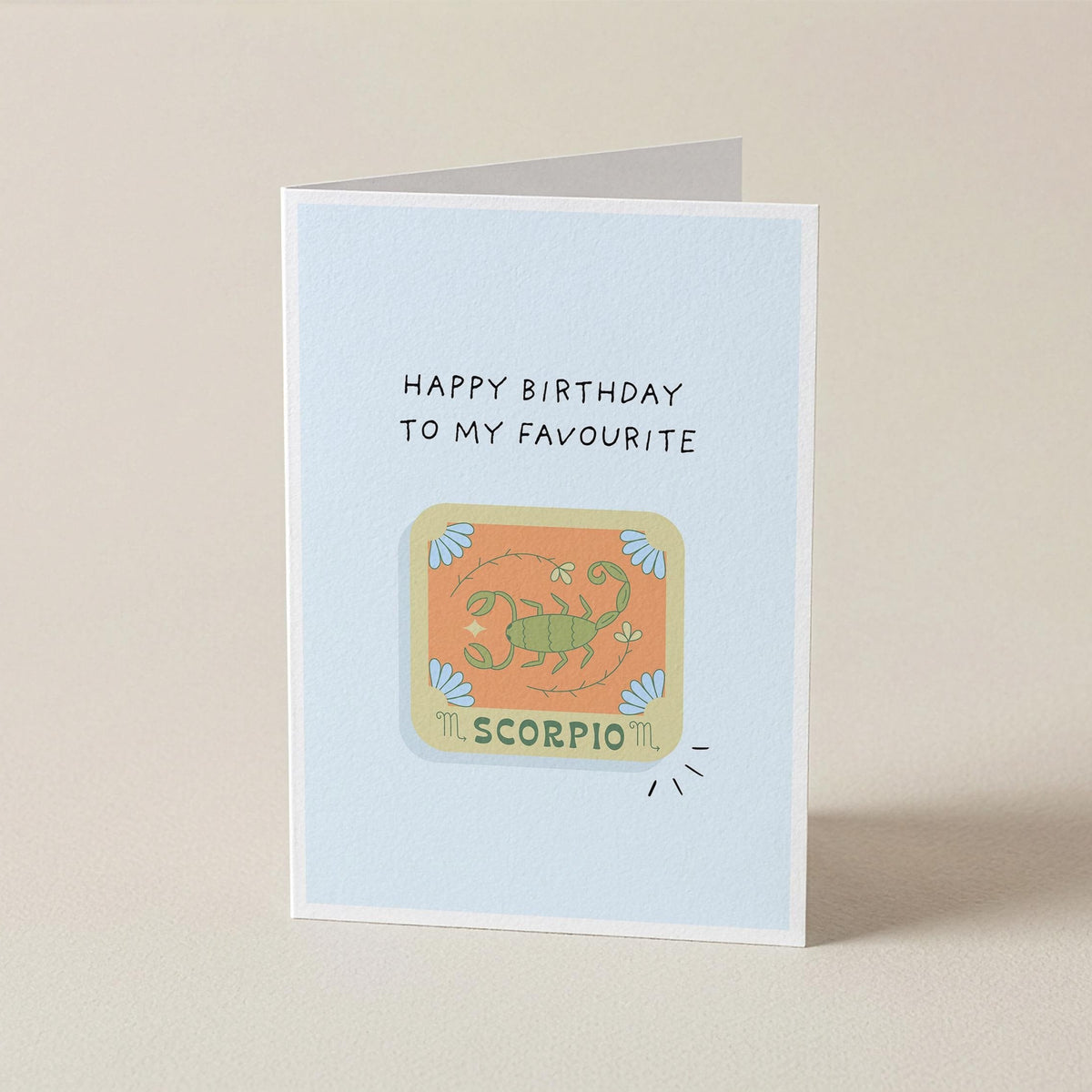 A folded greeting card standing upright on a light beige surface. The card has a pale blue front with the text 'HAPPY BIRTHDAY TO MY FAVORITE' above a colorful Scorpio illustration and the word 'SCORPIO'.